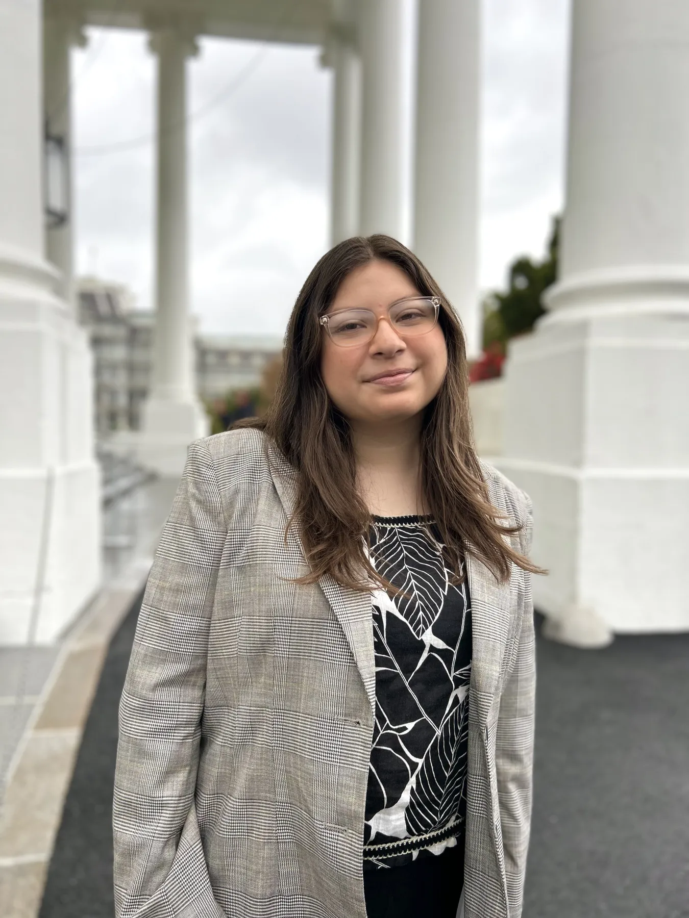 A photo of a woman with long brown hair and glasses outside a government building