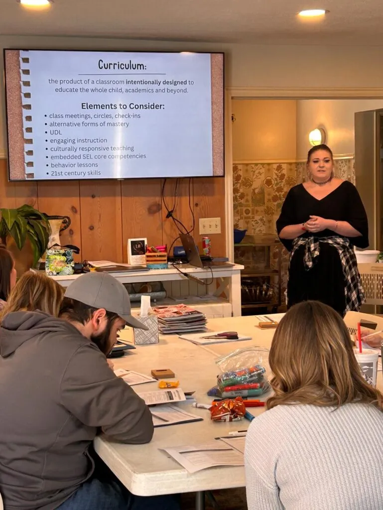 A woman dressed in black addressing a group of seated people. There is a video screen behind her with the heading Curriculum.