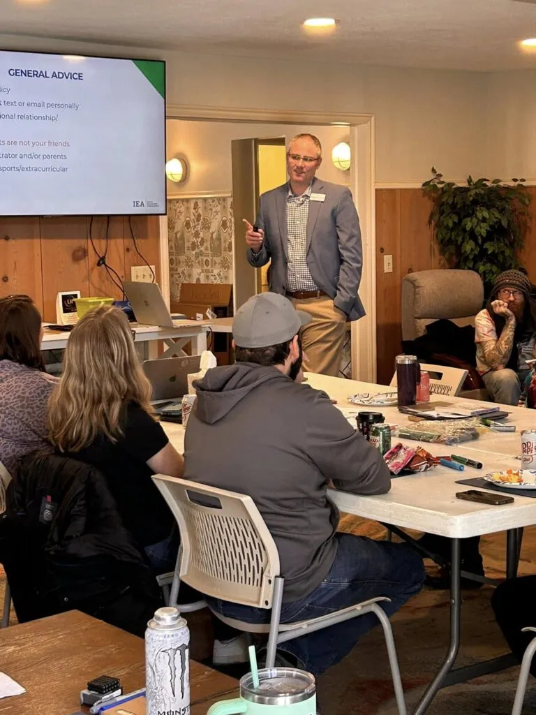 A man in a gray blazer gesturing as he speaks to a group of people seated around a table