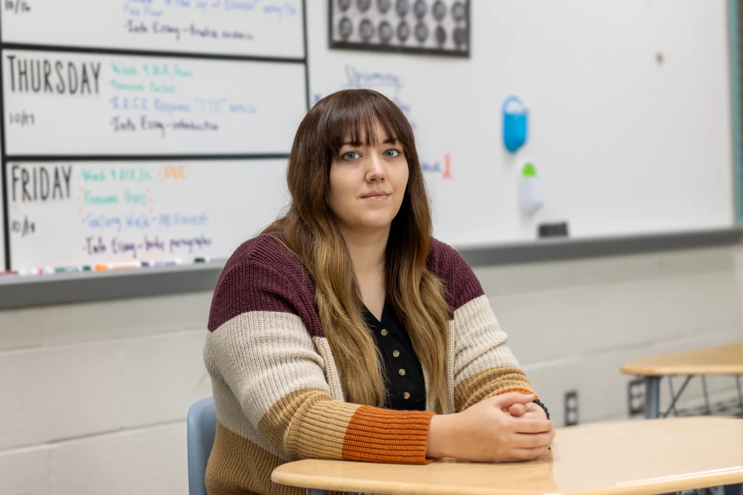Teacher sits at desk with a white board in the background.