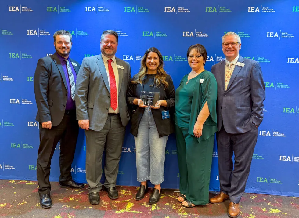 A woman in a black jacket and jeans is holding an award and surrounded by smiling people