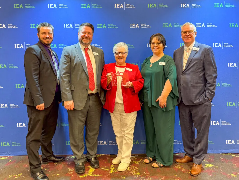 A woman in a red blazer and white pants holds an award while a group surrounds her