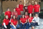 A group of smiling people wearing red shirts pose beneath a tree