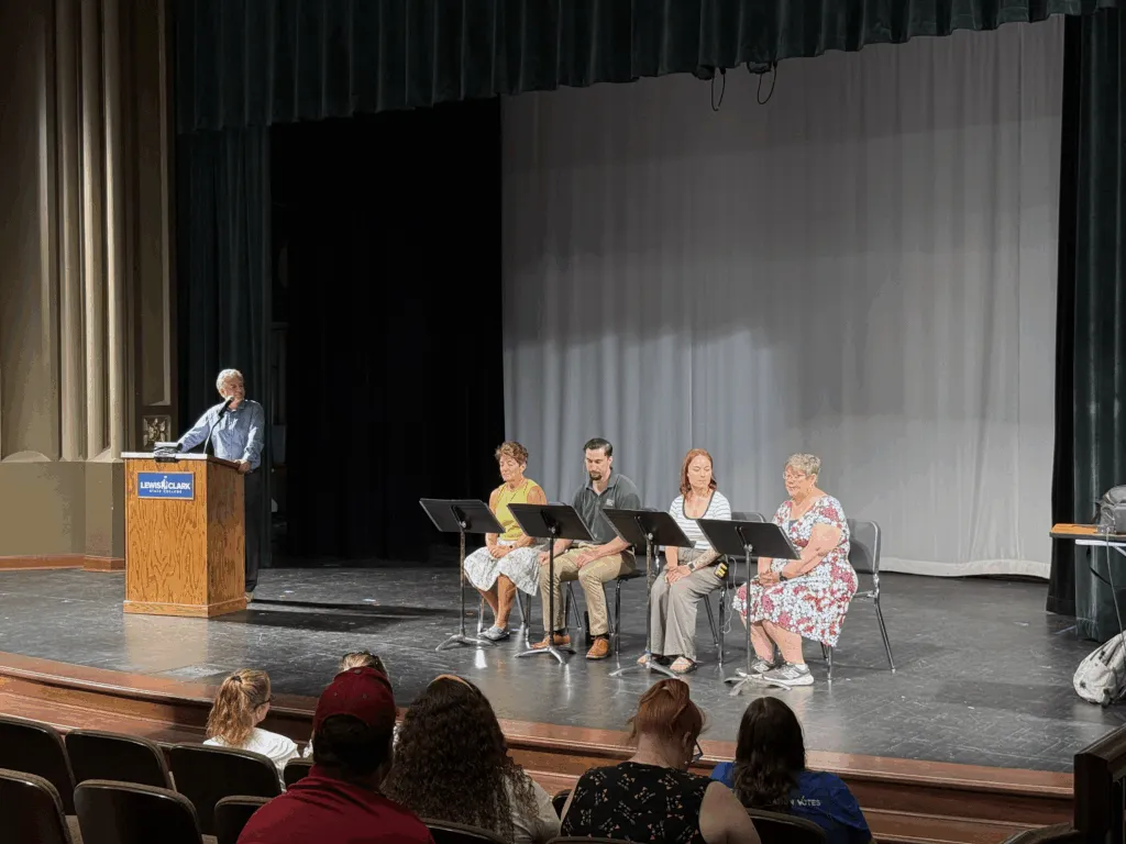 Four people sit on stage, reading, while a man looks at them from a podium. 