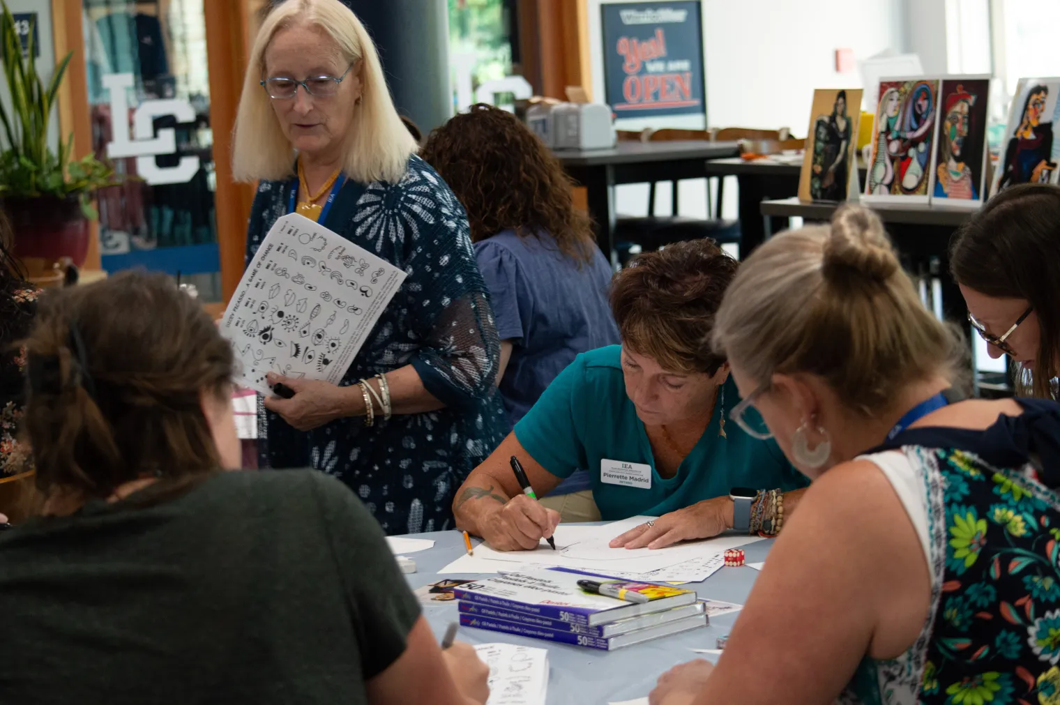 A woman looks over the shoulder of women creating art at a table. 