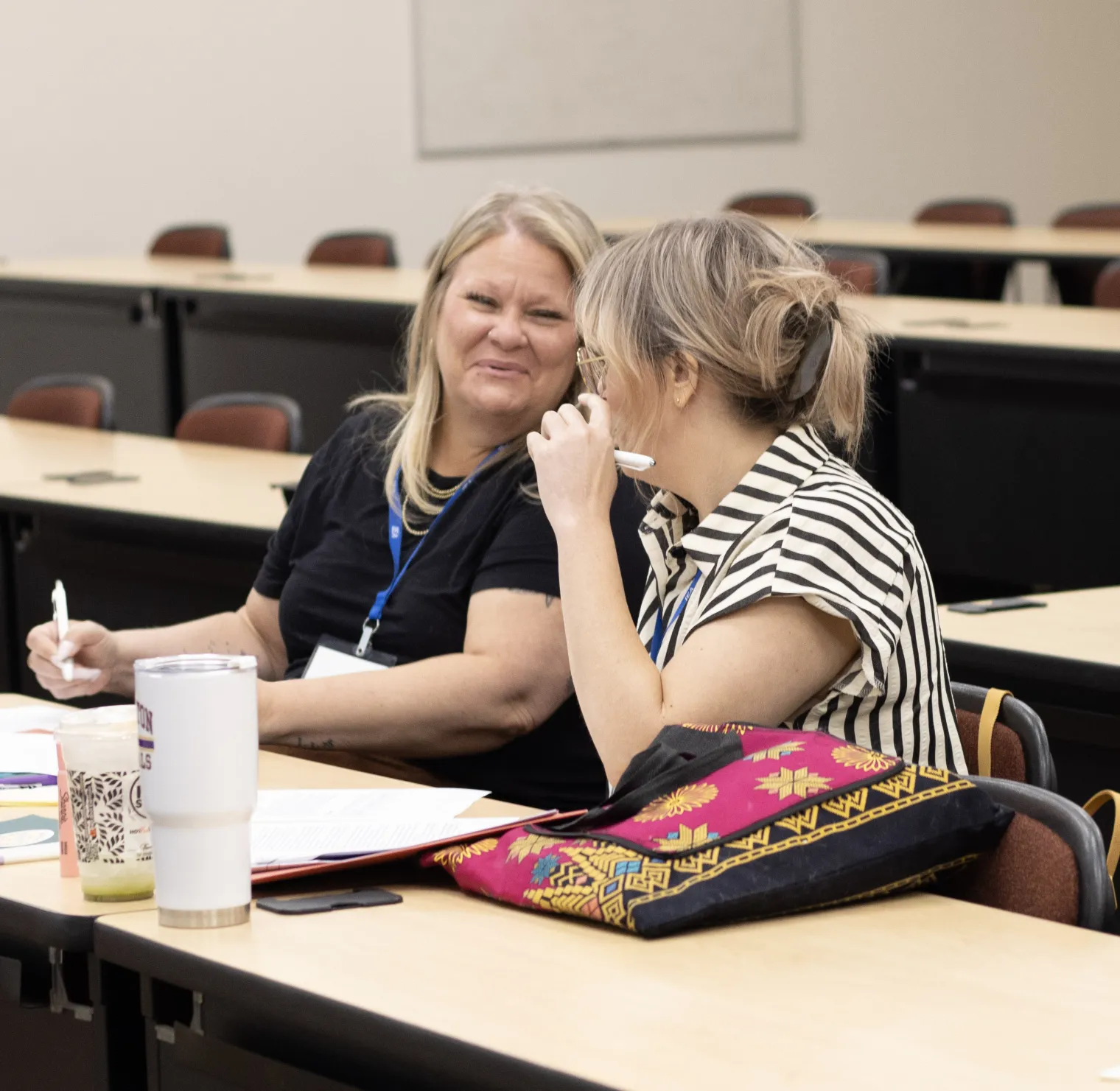 Two seated women in a classroom speak to each other