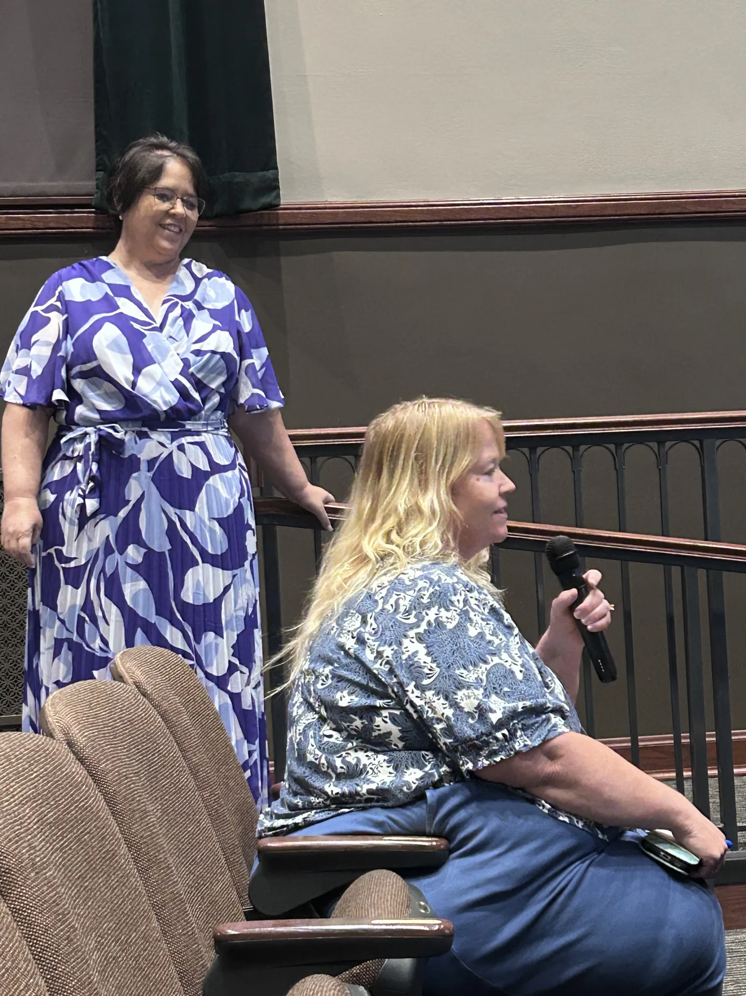 A woman standing next to a seated woman speaking into a microphone
