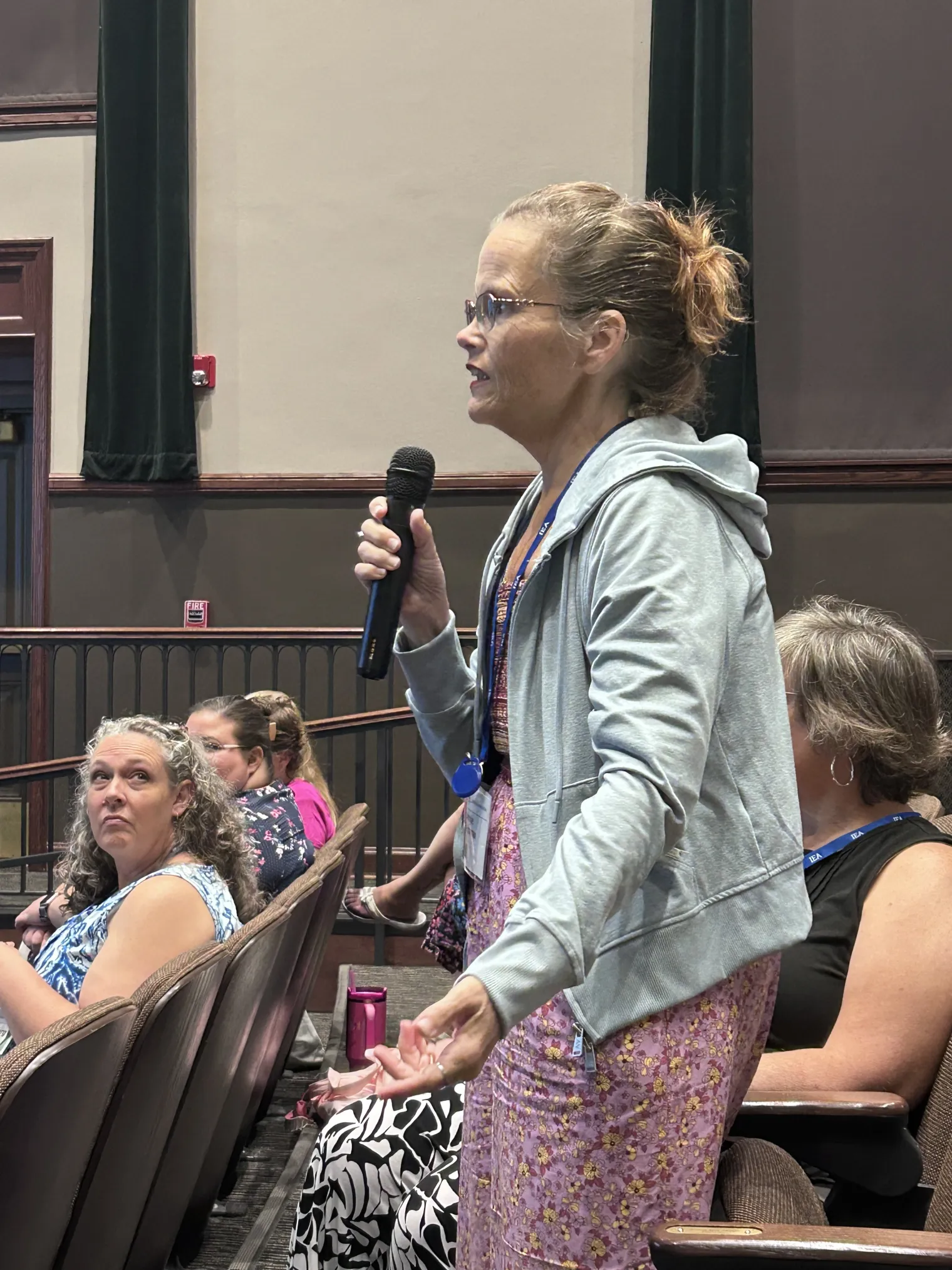 A woman with pulled back hair in a gray hoodie speaks into a microphone
