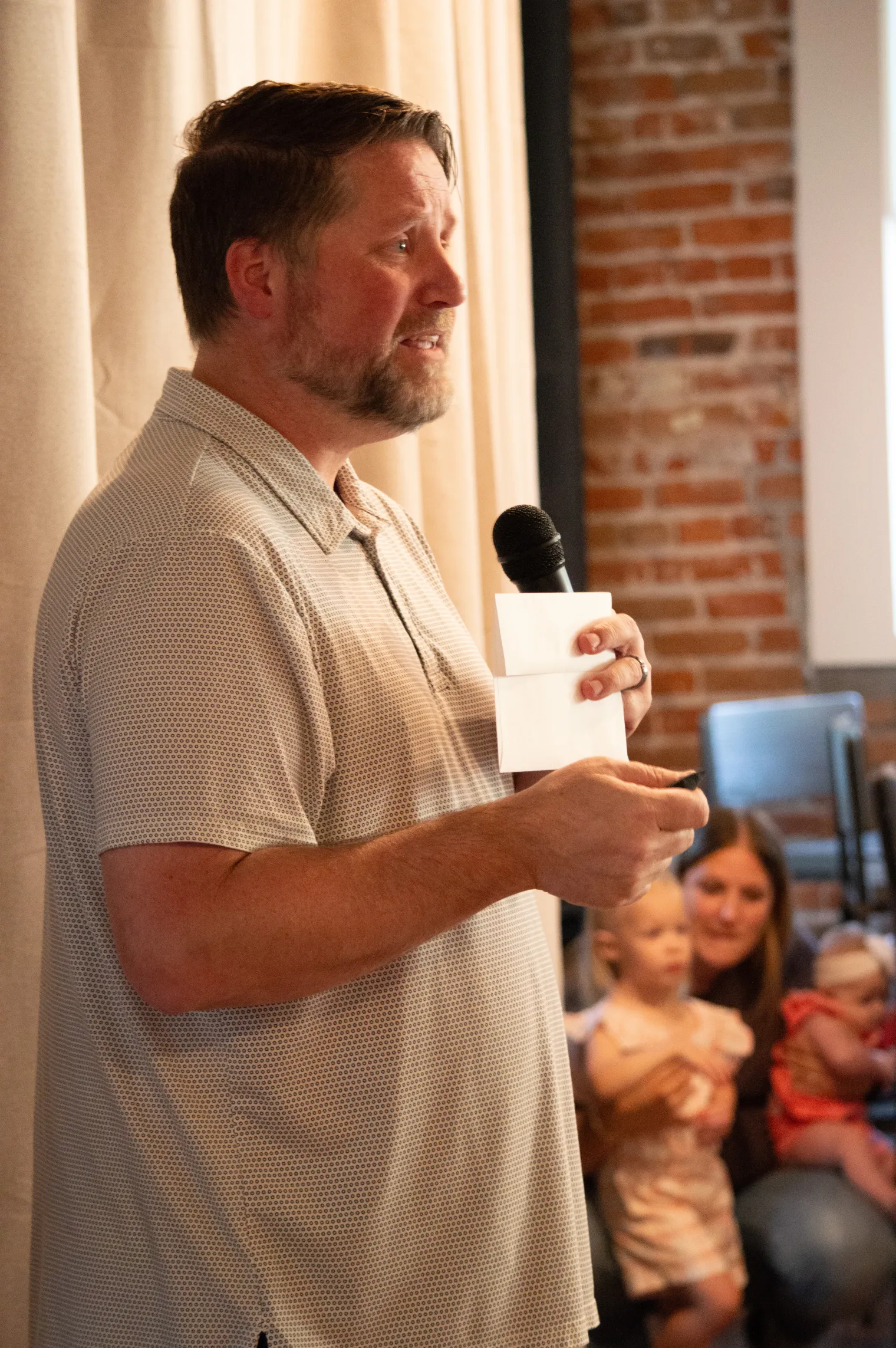 A man in a white polo shirt holds a microphone and speaks