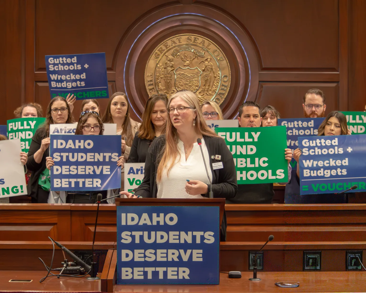 IEA member Melyssa Ferro speaks at a podium with group of teachers holding signs behind her.