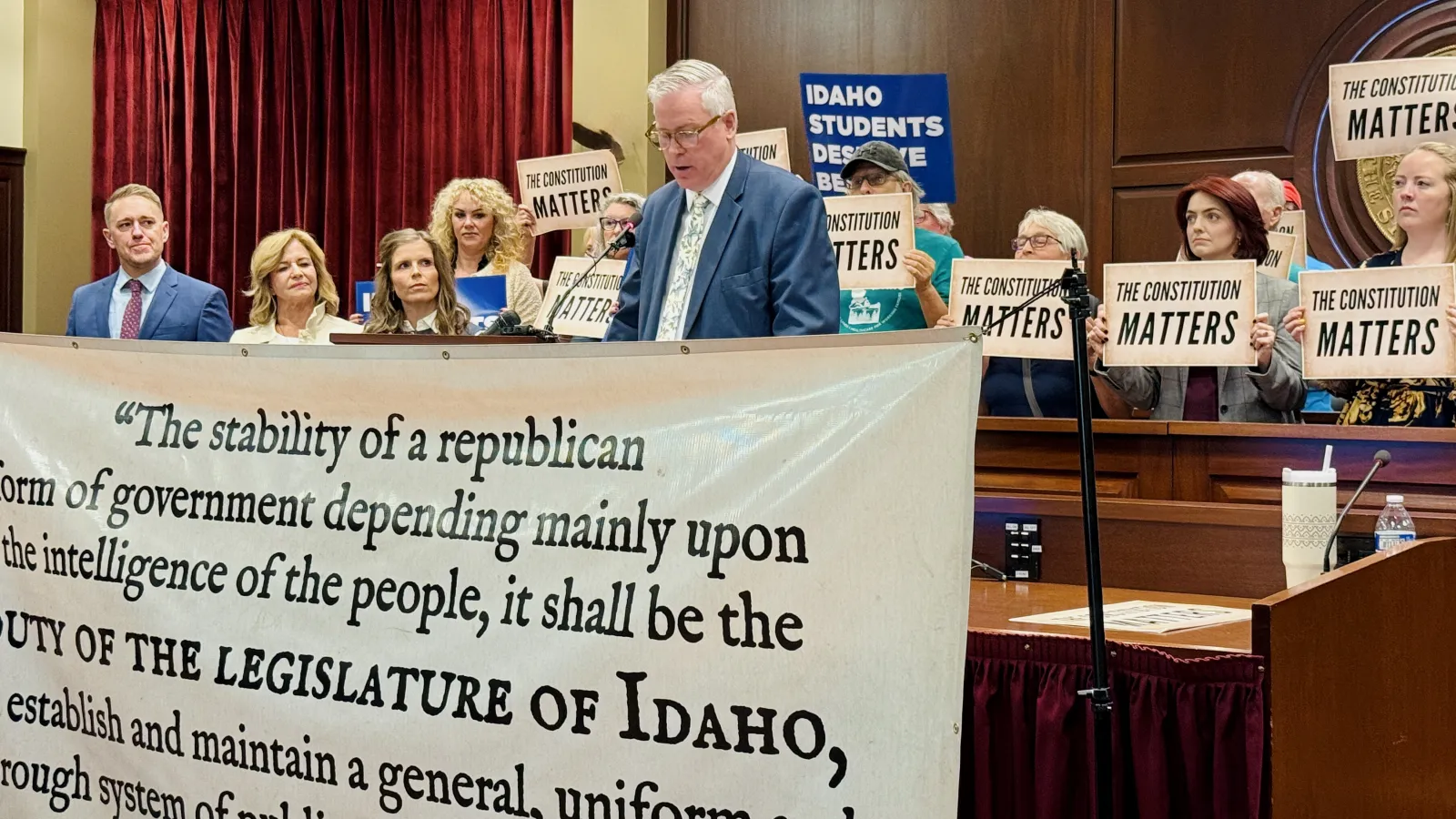 A man with white hair in a blue suit and tie reads remarks from a podium. He is surrounded by people holding signs. In front of him is a banner with a portion of the Idaho Constitution written on it. 