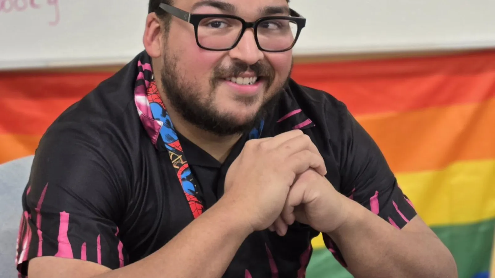 A bearded, bespectacled person poses at a desk in front of a Pride flag