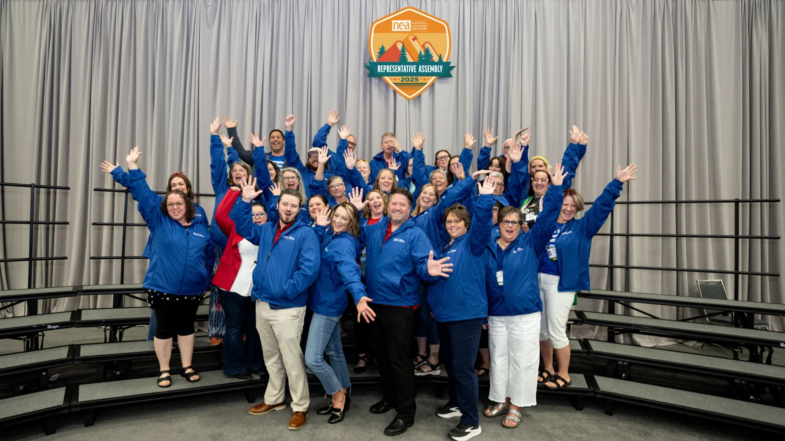 IEA's 2025 NEA Representative Assembly delegates pose for a delegation photo under the RA logo with their arms in the air, dressed all in blue.