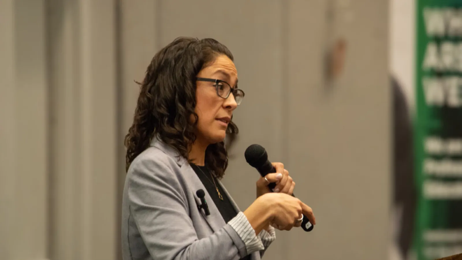 A dark haired woman wearing glasses and a gray blazer speaks into a microphone