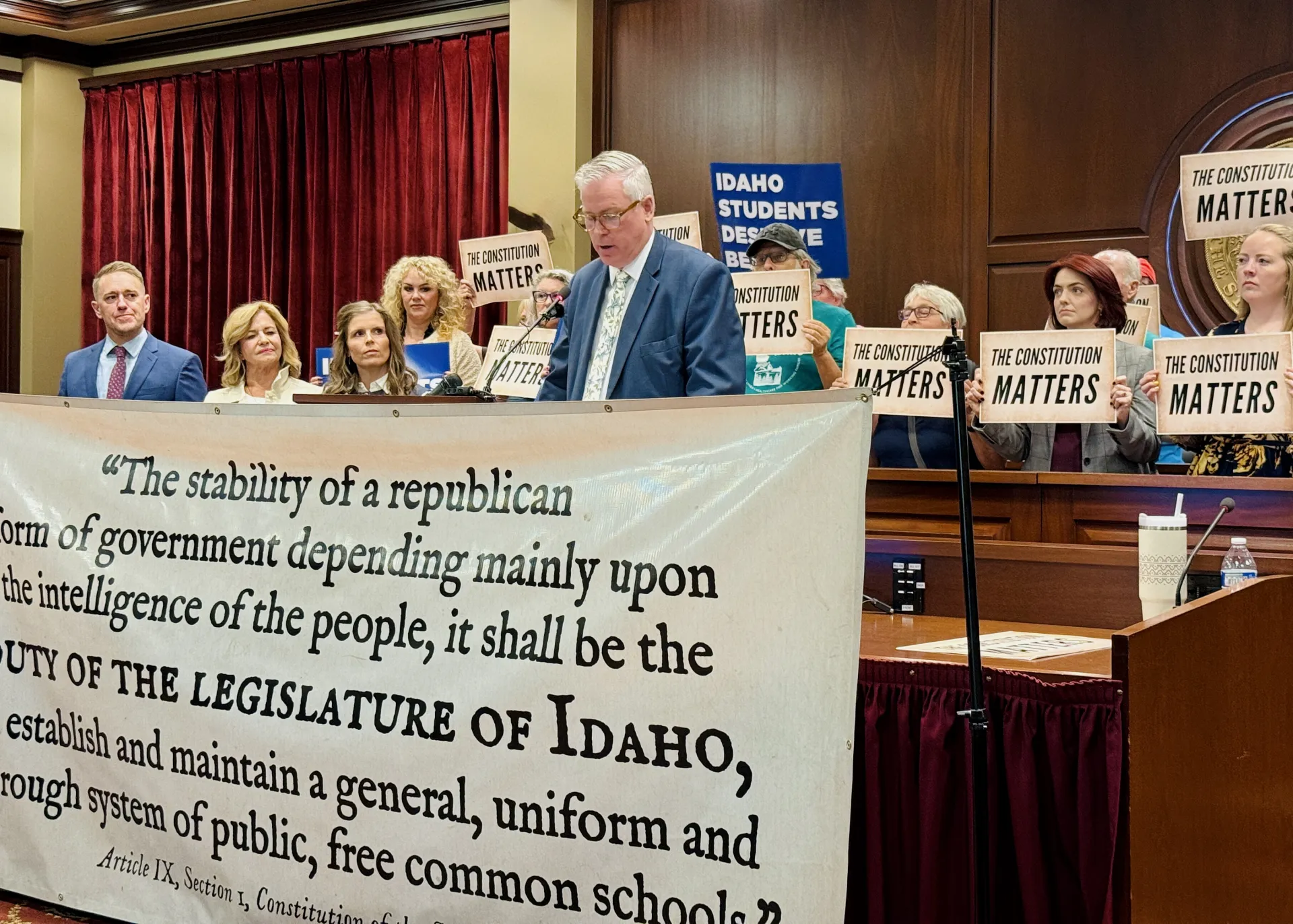 A man with white hair in a blue suit and tie reads remarks from a podium. He is surrounded by people holding signs. In front of him is a banner with a portion of the Idaho Constitution written on it. 