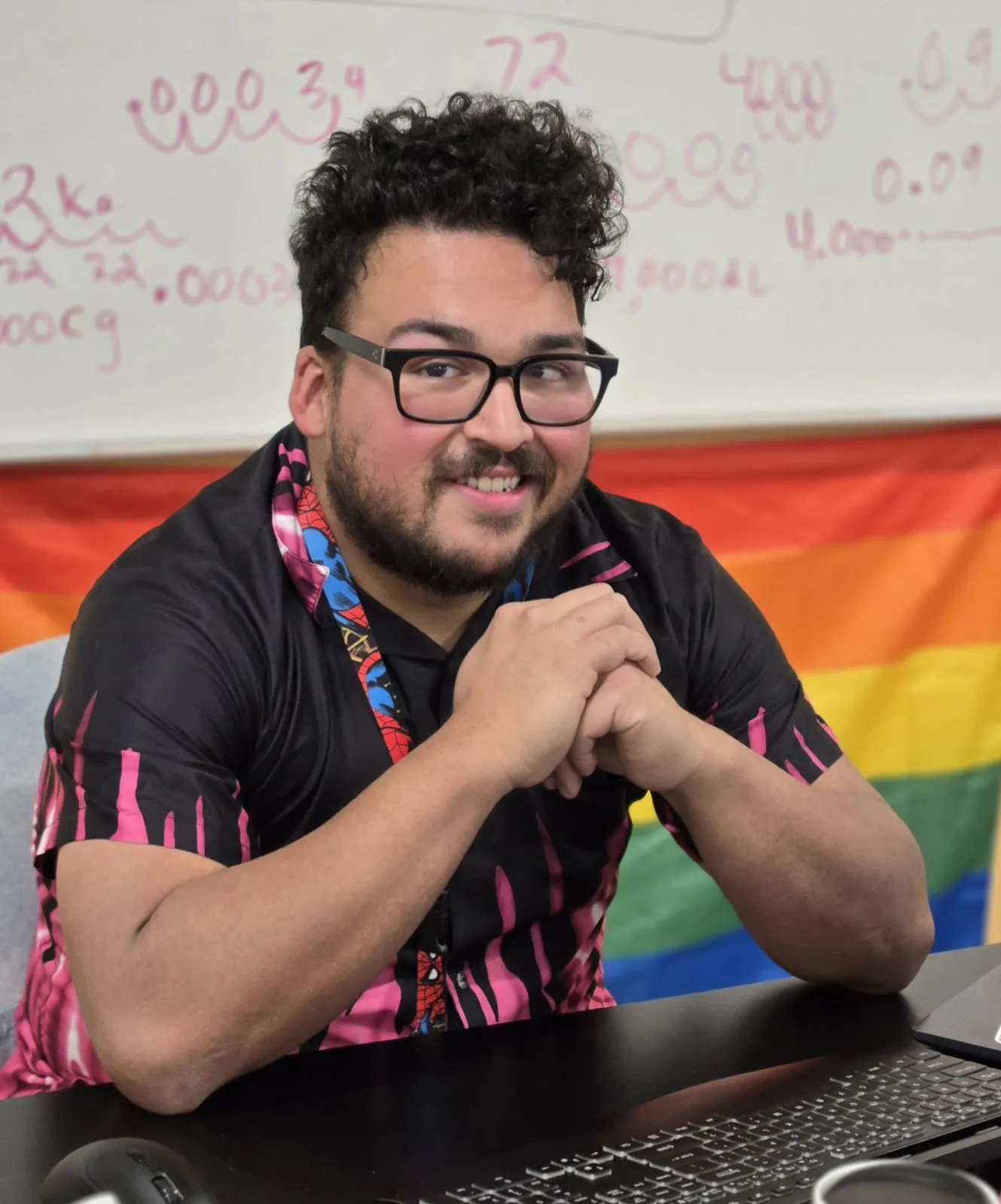 A bearded, bespectacled person poses at a desk in front of a Pride flag