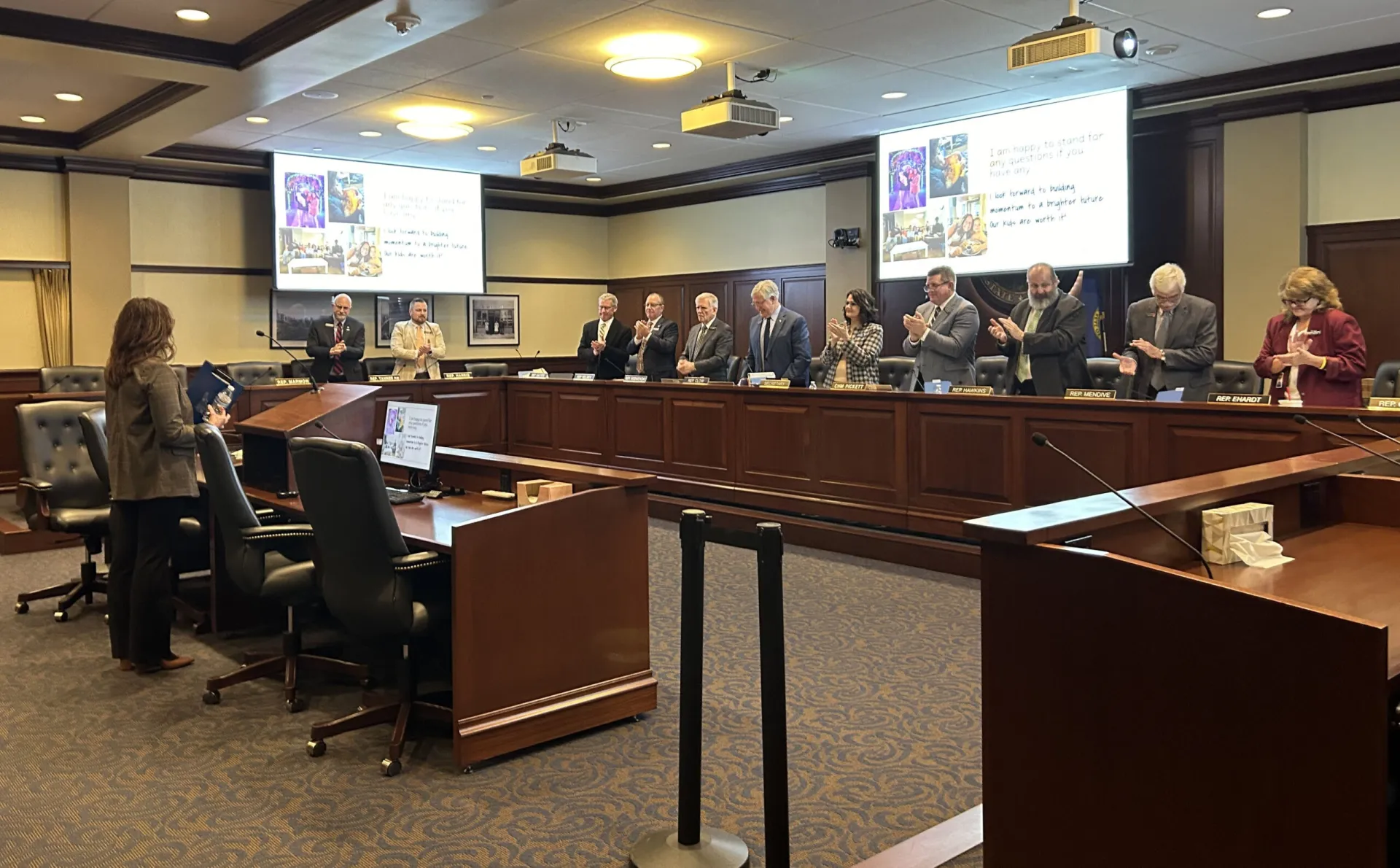 A woman stands at a podium in a legislative committee room while lawmakers give her a standing ovation 