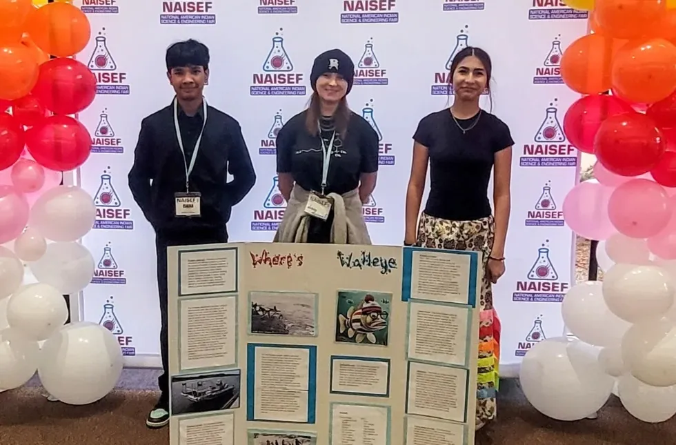 A group of three smiling students pose behind a science project