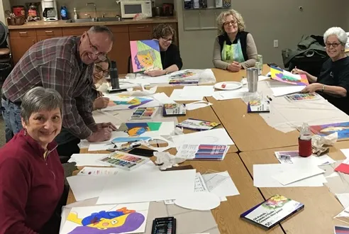 A group of people seated around a large table show off their art projects