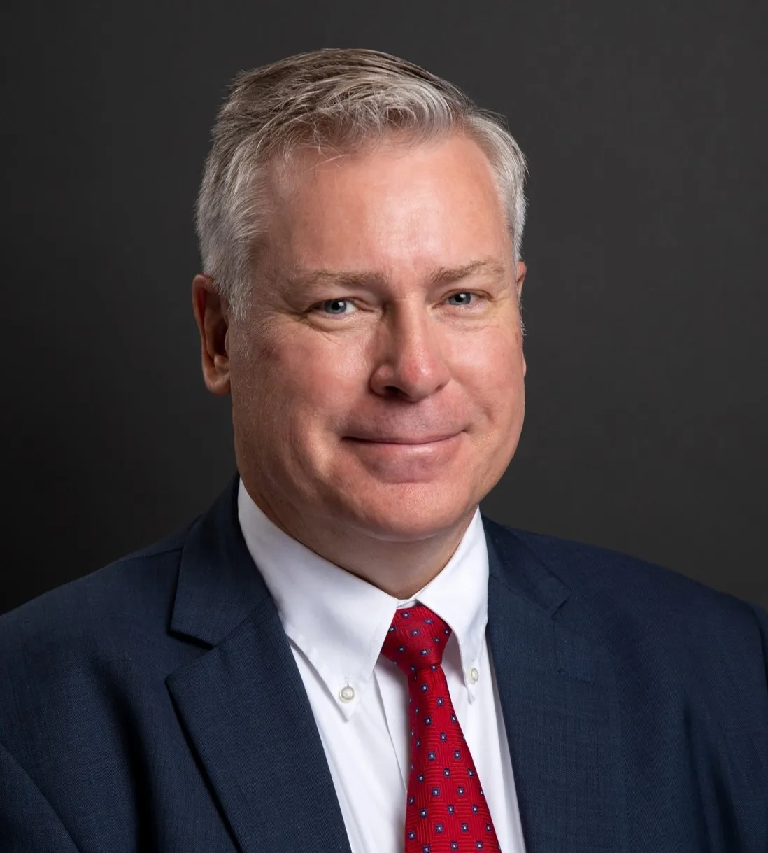 A man in a blue suit jacket and red tie smiles at the camera
