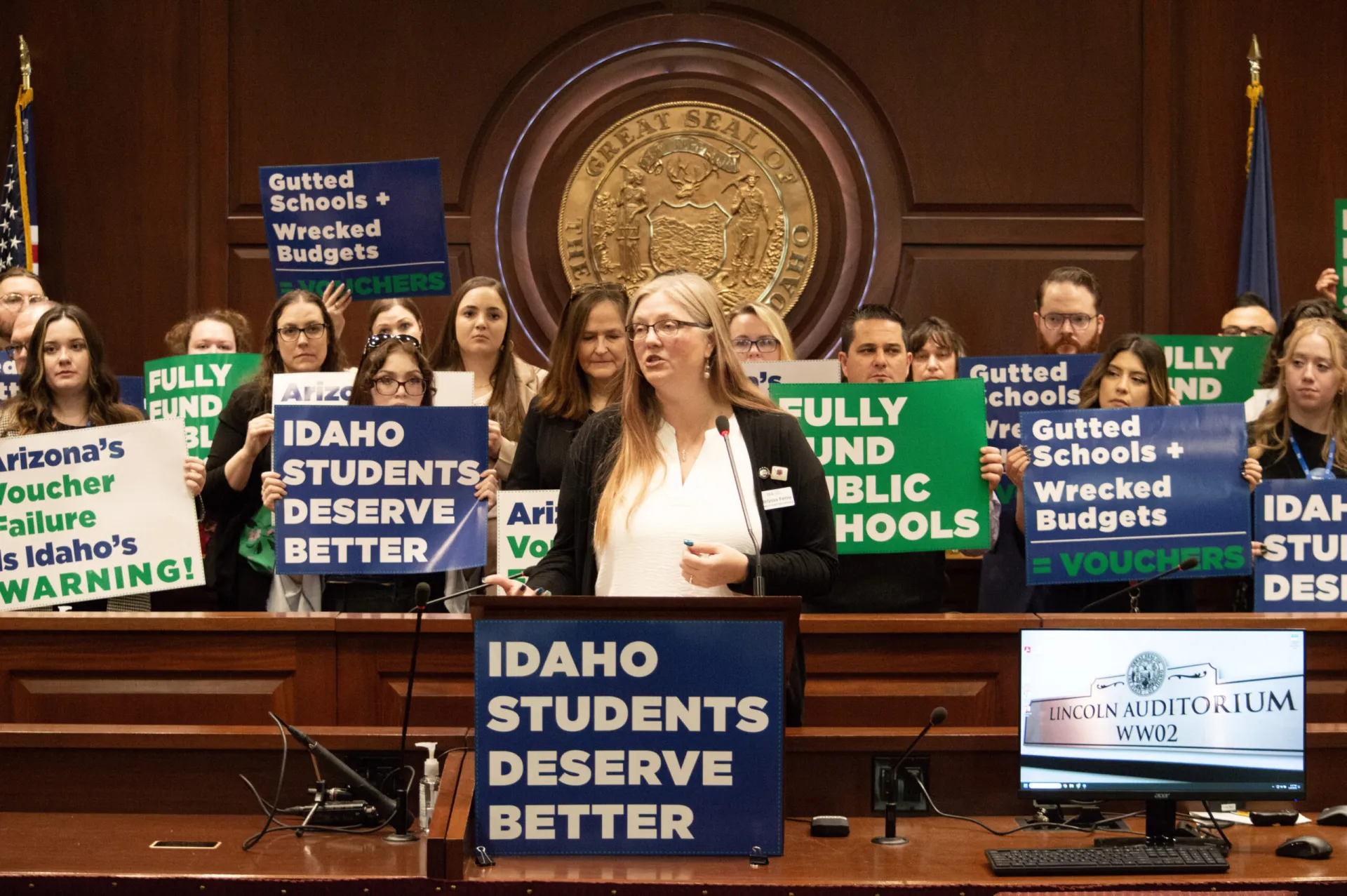 A woman speaks at a podium as a group of people hold blue, green and white anti-voucher posters