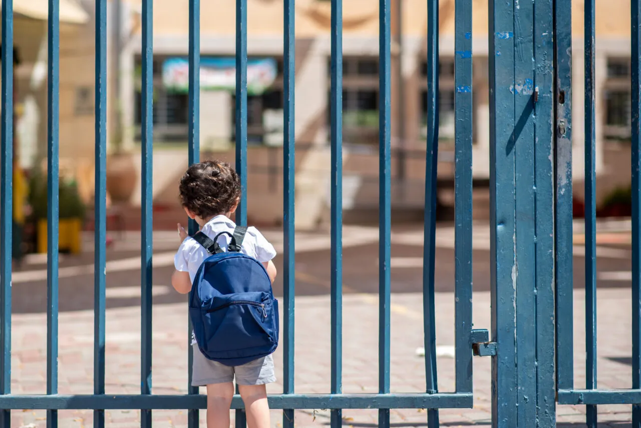Small child wearing a blue backpack stands at locked gate.