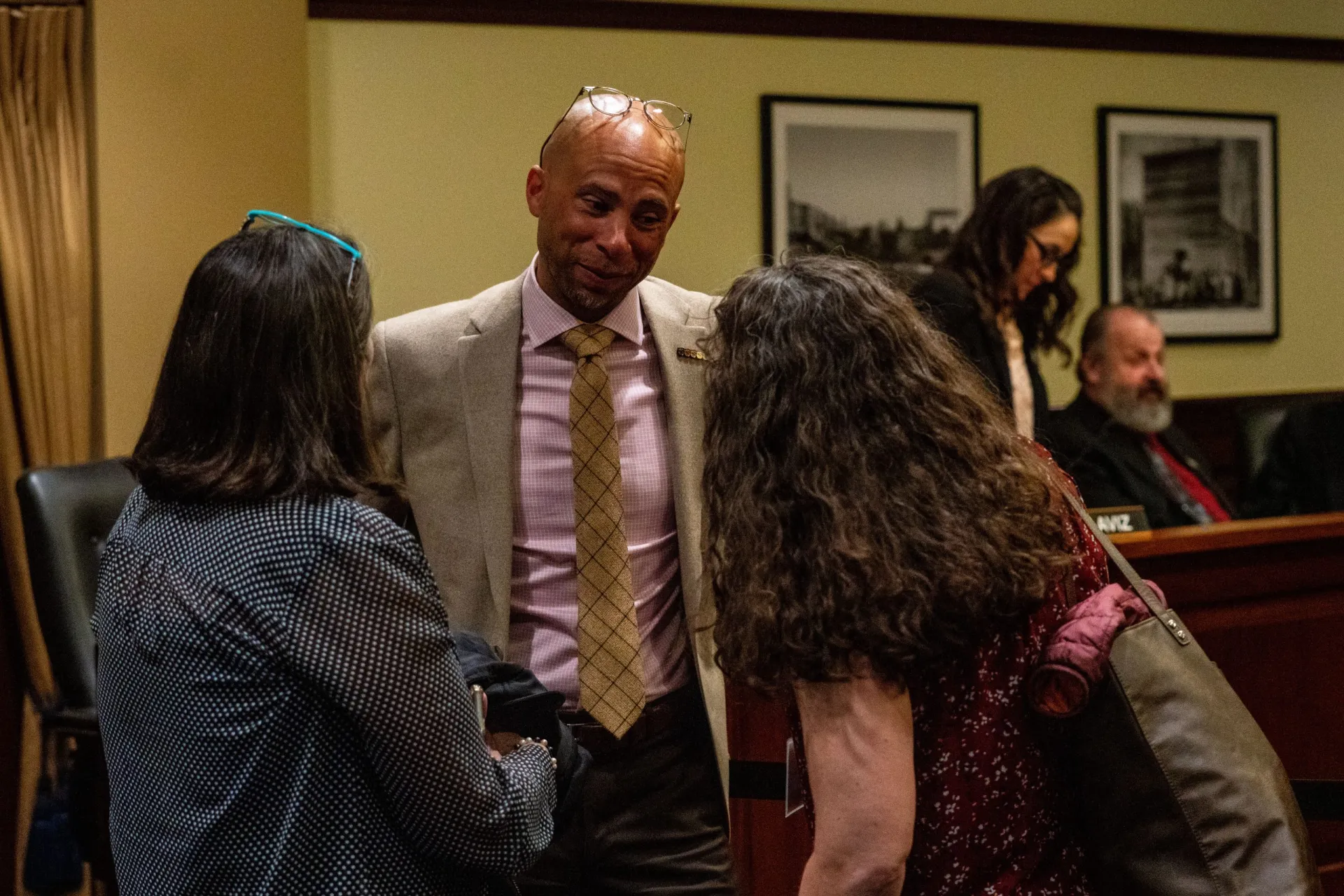 A bald man wearing a pink shirt and tan blazer and tie speaks to two women while smiling