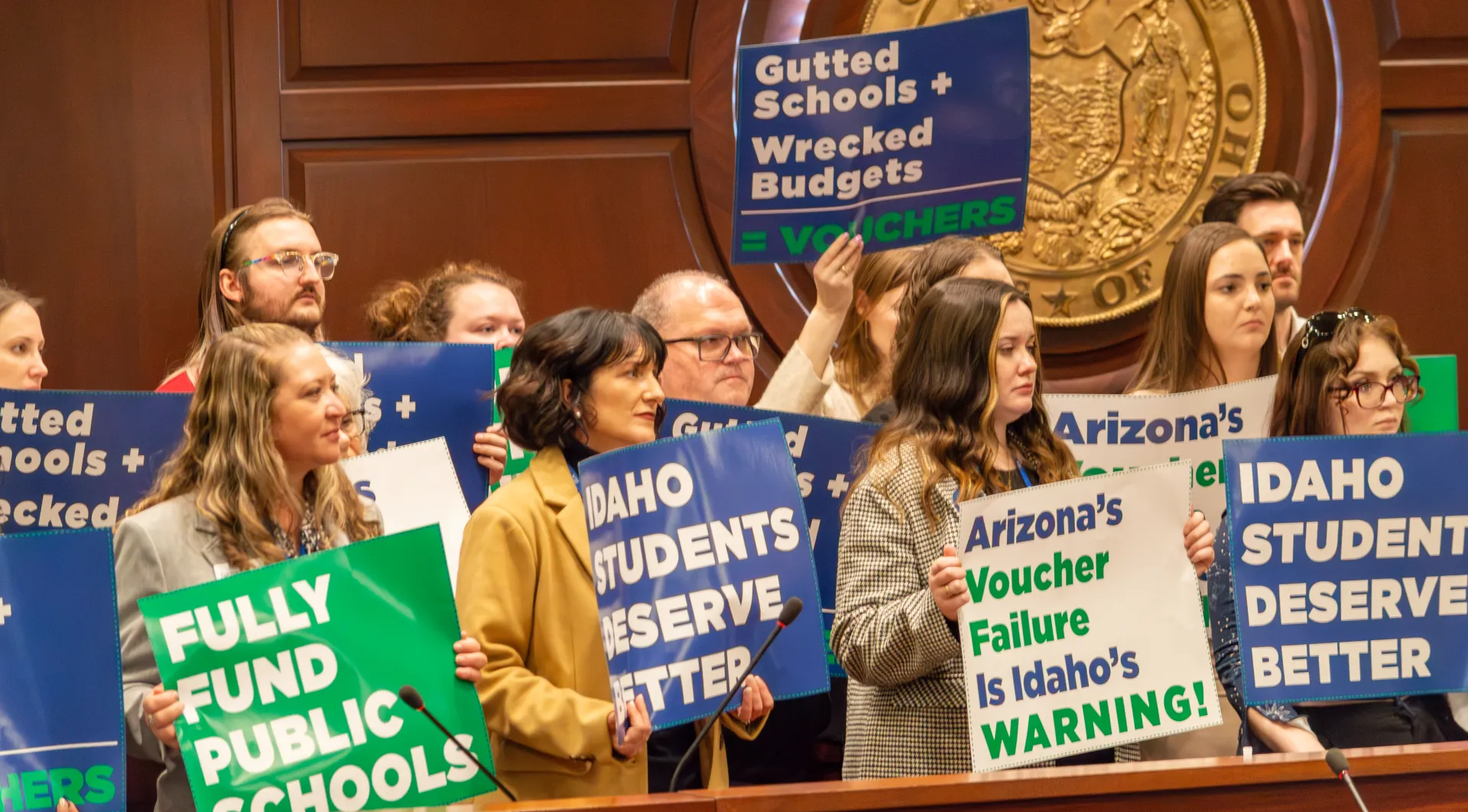 Group holding blue and green signs, listening to a speaker.
