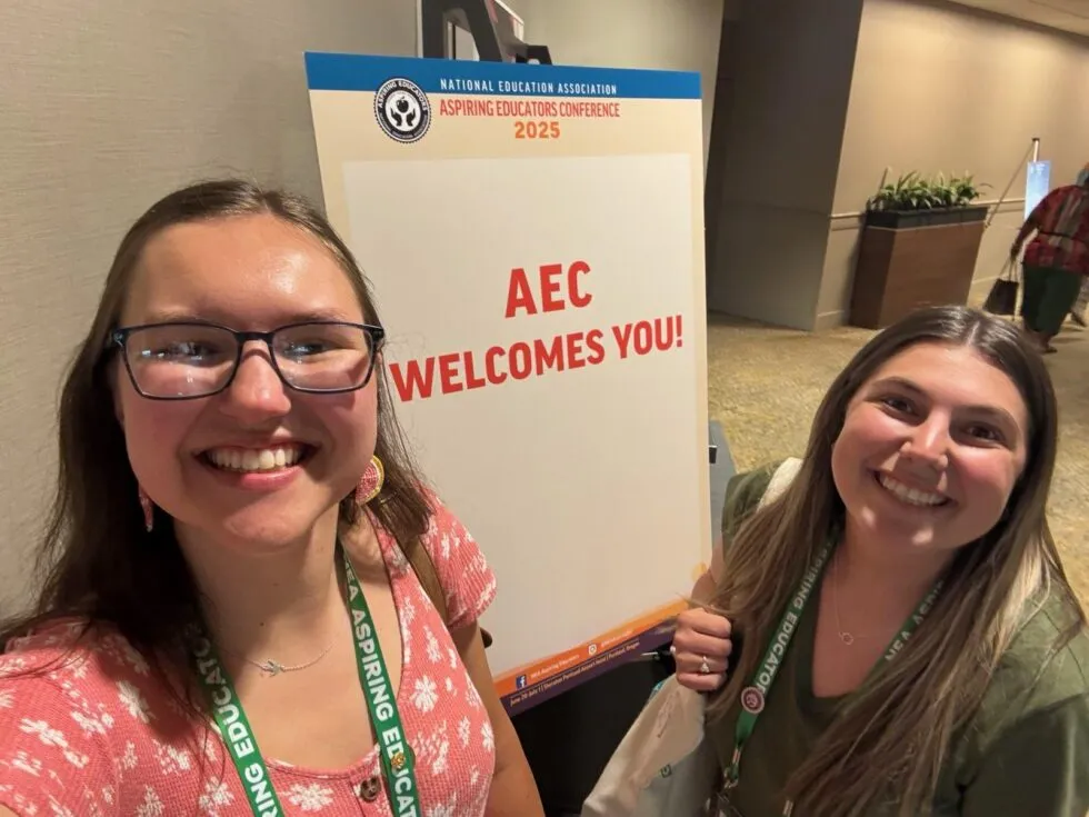 Two young women smile for a selfie before a poster that says AEC Welcomes You