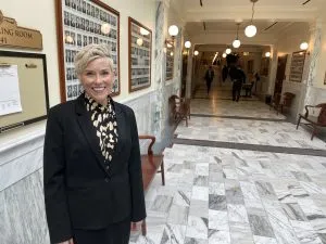 A woman with short blonde hair and wearing a black suit stands in an Idaho Statehouse corridor