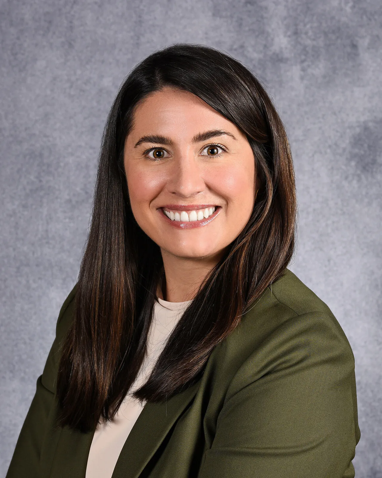 A woman with long brown hair wearing an olive colored blazer smiles at the camera