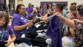 A woman in a purple shirt and glasses assists someone at a busy table piled full of clothes
