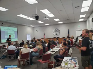 A group of people sitting at desks listen to a person speaking before a screen