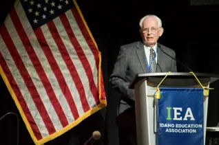 A man with white hair and glasses speaks at a podium
