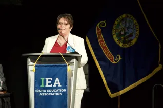 A woman in a white suit and red shirt speaks at a podium