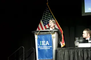 A woman in a black sweater and glasses speaks at a podium
