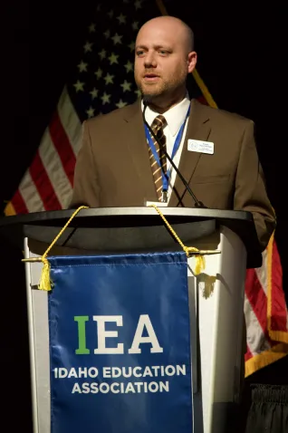 A man in a brown suit speaks at a podium