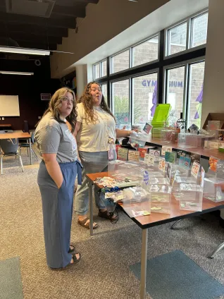 Two women stand over a table full of items and react with surprise to something someone said off camera