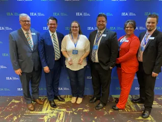 A woman in a white shirt and dark pants holds an award surrounded by IEA leadership