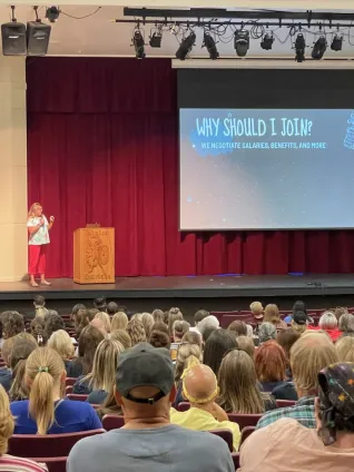 A photo of a woman on stage speaking to a crowd. The screen behind her says 'Why should I Join?"