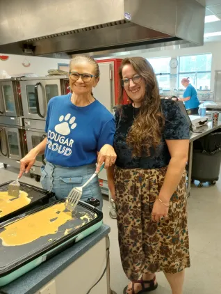 Two women in a cafeteria smile at the camera. The woman on the left is making eggs. 