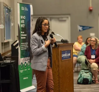 A woman wearing a gray blazer holds a mic and speaks to a group
