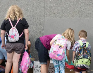 A group of children wear backpacks with their backs to the camera