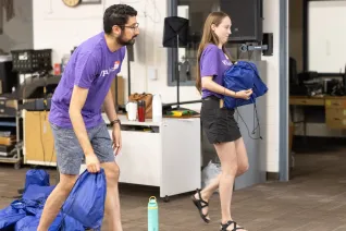 A man and a woman wearing purple shirts carry items