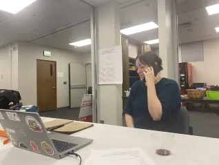 A woman seated at a table with a computer in front of her makes phone calls