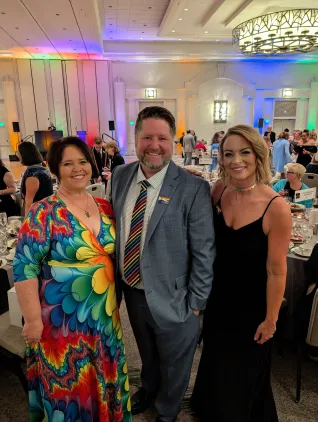 Two women and a man wear rainbow attire at a gala dinner