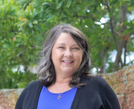 A smiling woman with salt-and-pepper hair wearing a purple shirt and black sweater