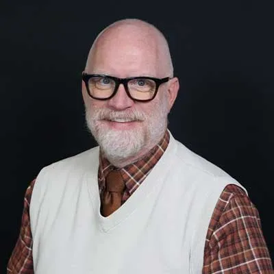 A man with a white beard and black glasses smiles at the camera