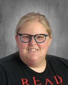 A woman with blonde hair and blue-green glasses smiles at the camera in a school picture