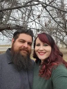 A man with a long dark beard and a woman with long red hair smile at the camera